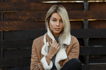 Outdoor portrait of a cute amazing young blonde girl with blue eyes and natural make-up on the background of a wooden vintage wall. Stylish autumn and spring outerwear. Attractive model woman