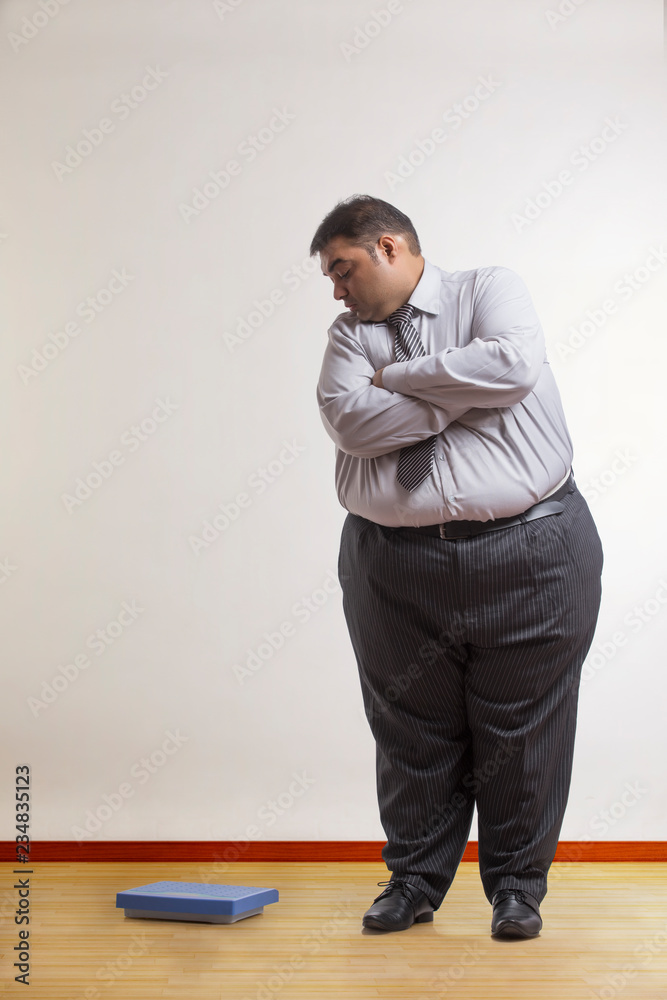 Obese man in formal clothes standing with arms crossed looking at weighing scale beside him Stock Photo | Adobe Stock Obese man in formal clothes standing with arms crossed looking at weighing scale beside him Stock Photo | Adobe Stock