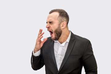 Sleepy young man in suit stands and yawns. He hold hand in front on mouth. Isolated on white background.