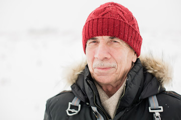 Close up portrait of  senior man standing outdoors in red hat and black jacket winter season. Snowy weather.