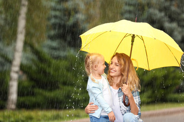 Wall Mural - Portrait of happy mother and daughter with yellow umbrella in park on rainy day. Space for text