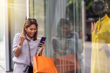 Asian woman cheerful when using the smart mobile phone for check online shopping order is completed with clothes beside the glassess in store shop with happy action at department center