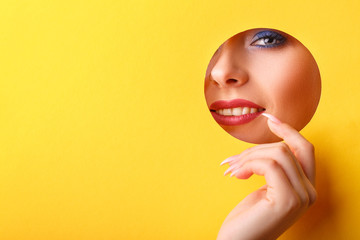 Woman looking in the hole, bright beautiful makeup, big eyes and lips, bright lipstick, professional cosmetics and facial care. Bright colored background and a gap slot in the paper