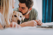 Pleasant bearded man kissing his dog while lying in bed with his girlfriend