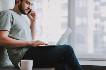 Young beared man talking on phone while using his laptop