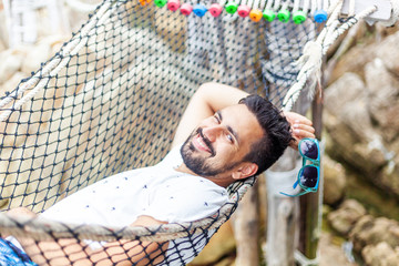 Wall Mural - Attractive handsome young latin brunette man in sunglasses with a beard on a hammock on vacation