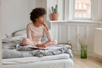 Pretty young dark skinned girl with Afro haircut, writes in diary, has dreamy expression, wears pyjamas, poses on bed in spacious bedroom, being deep in thoughts before sleep. Bedding concept