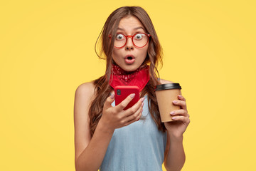 Wall Mural - Emotive surprised teenager girl opens mouth, feels amazement, holds smart phone and take out coffee, has impressed stupefied gaze, recieves unexpected message, isolated over yellow background