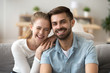 © fizkes - Portrait of smiling young husband and wife hugging sitting on couch, happy millennial couple look at camera posing for family picture at home, laughing man and woman hug having fun together