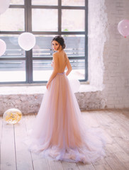 A graceful young lady with dark hair in a gorgeous tender long peach and purple dress with a train and open back, turns to the camera in a bright and spacious room filled with light color balls