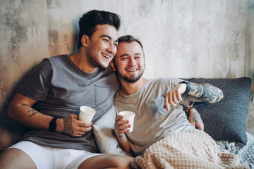 Wall Mural - Perfect day starts with sweetheart and coffee in bed. Two handsome gay men in bed resting and having morning cups of hot drink. They both engaged in video game, too far from urban fuss.