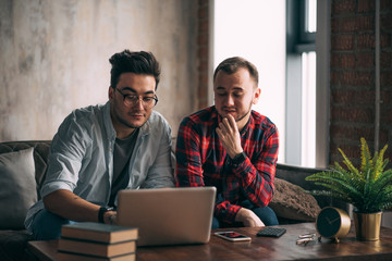 Wall Mural - Romantic partners in love and business. Caucasian amused gay couple looking at laptop screen while resting in living room with loft interior. Dominant man wearing spectacles and and using laptop