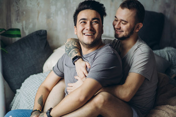 Wall Mural - Happy homosexual couple cuddling after a big quarrel, hugging during sexual foreplay. Handsome man with tattooed arm embracing his partner and smiling, sitting on bed in lounge monochrome interior.