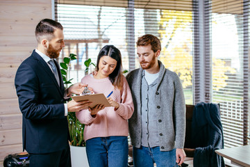 People, business and real estate. Young couple excited by luxury apartment, proposed for rent. Handsome caucasian realtor suggests to clients to sign rent property contract