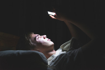 Depressed teenager browsing the internet on his mobile phone as he is lying on his bed in the dark.