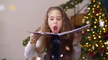 Portrait Of Adorable Preadolescent Girl Blowing Multi Colored Glitter Confetti From Open Hardcover Book Over Christmas Decorated Room Background. Beautiful Child Having Fun With Shiny Confetti At Xmas