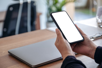 Mockup image of hands holding black mobile phone with blank white screen with laptop on wooden table