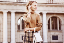 Outdoor Fashion Portrait Of Young Happy Smiling Lady Wearing Trendy Beige Turtleneck, High-waisted Checkered Trousers, Wrist Watch, Belt, Carrying Small White Bag, Posing In Street. Copy, Empty Space