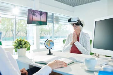 Cheerful excited senior lady in white cardigan sitting at table and keeping hands together while watching 3D video of hotel presentation with awe