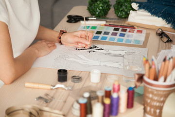 Wall Mural - Female clothing designer sketching, drawing with brush at workplace close up in cozy workshop studio, fashion designer, dressmaker creating new fashionable handmade clothes collection, hands view