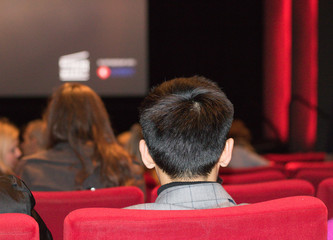 Man with a short hairstyle is sitting in a cinema chair on a blurred background of big screen and audience. It`s before a film starting. Concept: going to a cinema.