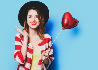 Portrait of young smiling red-haired white european woman in hat and red striped shirt with heart shape toy on blue background