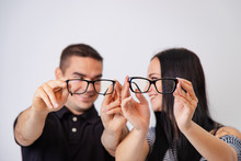 Lovely Attractive Couple Sitting Together And Looking Each Other Hold Glasses In Their Hands. Young Couple Smiling To One Another Show Eyeglasses In Black Frame Sitting On White Background. Close-up