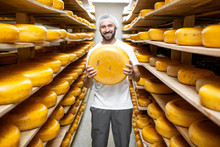 Portrait Of A Handsome Worker Standing With Cheese At The Storage Full Of Cheese Wheels During The Aging Process