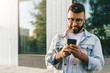 © foxyburrow - Handsome smiling hipster businessman with beard, in denim jacket, trendy glasses walks around city and using smartphone.