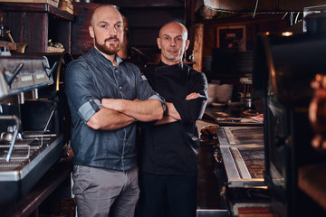 Chef with his assistant posing for a camera with his arms crossed in a restaurant kitchen.