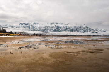 Calm reflective water sits still below cloud covered snowy mountain peaks