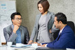 © DragonImages - Young businesswoman standing near the table and listening how her colleagues discussing business ideas at meeting at boardroom