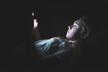 Depressed Girl Lying Down On A Couch In The Dark While Using Her Smartphone. The Light From The Screen Is Illuminating Her Face.