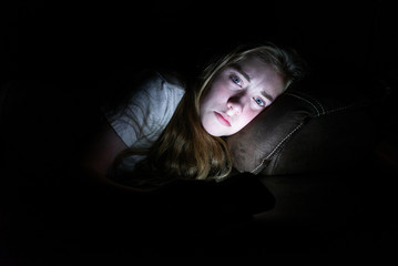 Depressed girl lying down on a couch in the dark while using her smartphone. The light from the screen is illuminating her face.