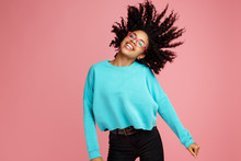 Excited African American Young Woman With Bright Smile Dressed In Casual Clothes, Glasses And Headphones Dance Over Pink Background.