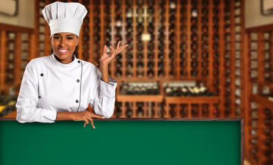 Black Brazilian woman chef cooking looking at camera with green board in wine house blurred background.