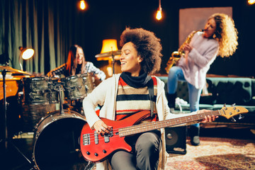 Girls playing jazz music. In foreground one woman playing bass guitar and in background other two playing saxophone and drums. Home studio interior.