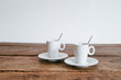 2 Espresso Tassen mit Löffel stehend auf Unterteller Holztisch Hintergrund weiß