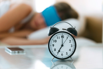 Wall Mural - Beautiful young sleepy woman with sleep mask resting in bed with alarm clock in bedroom at home.