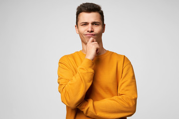 Wall Mural - Young man stands holding his hand on a chin, one corner of his lips is slightly pressed, looks thoughtfully, serious, thinking over a problem,with dissatisfied facial expression, over white background