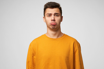 Wall Mural - Expresses insult childly. Portrait of emotional guy looks sad, upset, frustrated, displeased, lower lip pouted, showing defenseless, wears orange sweatshirt, stands isolated over white background.