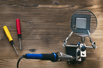 Retro computer CPU chip through magnifying glass mounted on the soldering stand and soldering iron on technician engineer table background with copy space.