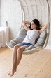 © fizkes - Smiling young barefoot woman sitting, relaxing with hands behind head in lounge hanging chair, looking in distance, dreaming about good future, happy girl resting in cozy, comfortable sofa at home