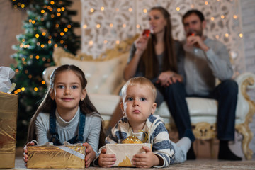 Brother and sister with gifts sitting on the floor against their parents and Christmas tree