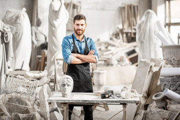 Portrait of a handsome sculptor in blue t-shirt and apron working with stone sculptures on the table at the old atmospheric studio