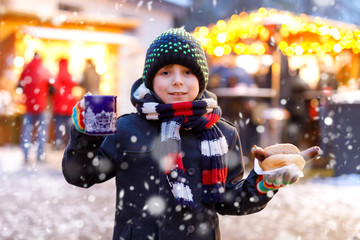 Wall Mural - Little cute kid boy eating German sausage and drinking hot children punch on Christmas market. Happy child on traditional family market in Germany, Munich. Laughing boy in colorful winter clothes