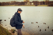Handsome Dark-haired Middle-aged Man In Jacket, Cap And Backpack Feeds Ducks On Pond In City Park In Late Autumn. Authentic Lifestyle Moments