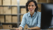 © Gorodenkoff - Female Inventory Manager Works Sitting at Her Desk, Working on Computer and Smiling at the Camera. In the Background Warehouse Storeroom with Shelves Full of Cardboard Box Packages Ready For Shipping