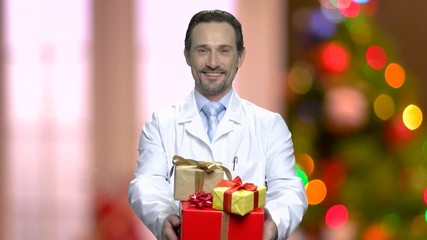 Wall Mural - Smiling doctor giving gifts on Christrmas background. Health care worker presents gifts boxes on defocused Christmas lights. Merry Christmas and Happy New Year.