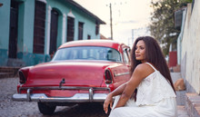 Beautiful Young Black Skinned Woman In A White Dress Sitting Besides An Old Classic Car At Sunset In The Streets Of Old Trinidad In Cuba.